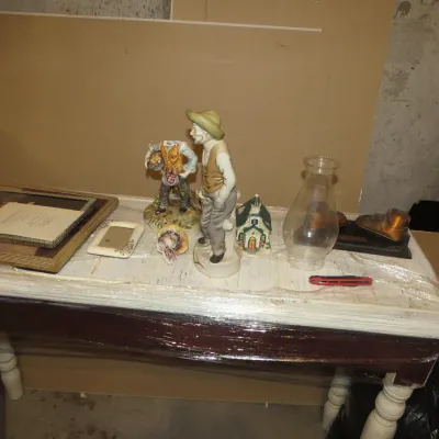 Sale Photo Thumbnail #58: Collection of vintage decor items, including ceramic figurines, framed pictures, a clear glass hurricane lamp chimney, and a small ceramic house model, displayed on a wooden table.