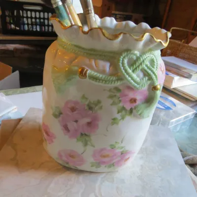Sale Photo Thumbnail #65: Hand-painted porcelain jar featuring pink floral motifs, a ruffled gold-rimmed top, and molded green cord decoration, holding an assortment of art paintbrushes.