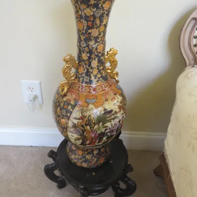 Sale Photo Thumbnail #9: Large Japanese Satsuma-style porcelain floor vase with gilt dragon handles, intricate narrative scene panels, and a ruffled rim, displayed on a carved dark wood stand.