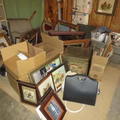 Sale Photo Thumbnail #55: Collection of framed artwork, empty picture frames, cardboard packing boxes, and storage items arranged in a basement or garage setting.