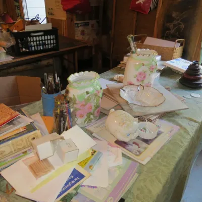 Sale Photo Thumbnail #54: Craft table filled with supplies including paintbrushes in jars, floral porcelain vases, paper pads, art prints, and a cat figurine.