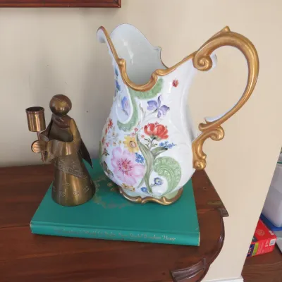 Sale Photo Thumbnail #15: Ornate hand-painted porcelain pitcher with floral motifs and gold scroll handle, alongside a vintage brass angel candlestick resting on a hardcover book.