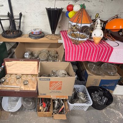 Sale Photo Thumbnail #88: Collection of vintage household items including an orange dome desk lamp, a stained glass style pendant light, glassware set in a wooden box, and various storage boxes filled with kitchenware and tools.