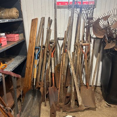 Sale Photo Thumbnail #53: Large assortment of vintage hand tools including shovels, rakes, pickaxes, and garden implements leaning against a wall.