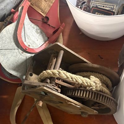 Sale Photo Thumbnail #67: Vintage heavy-duty hand winch with coiled rope, alongside an assortment of workshop gear including a red metal shield and a bucket of miscellaneous small items.