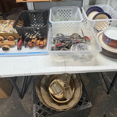 Sale Photo Thumbnail #91: Assortment of household goods on a folding table, including vintage wooden toy vehicles, kitchen utensils in plastic bins, ceramic plates, and lampshades underneath.
