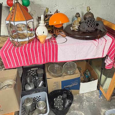 Sale Photo Thumbnail #89: Collection of vintage lamps and home decor, including an orange stained-glass pendant, a duck head lamp, and an orange mushroom lamp, displayed alongside boxes of glassware and aluminum kitchenware.