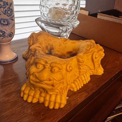 Sale Photo Thumbnail #39: Carved yellow resin or composite ashtray featuring ornate mythical lion or dragon face details, sitting on a wooden tabletop next to clear glass items and a hand-painted vase.