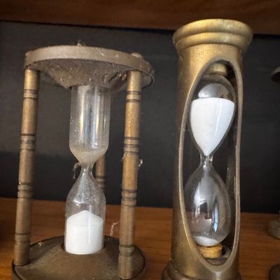 Sale Photo Thumbnail #43: Pair of vintage brass hourglasses, featuring one three-pillar style timer and one cylindrical cut-out design, both with white sand inside.