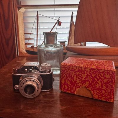Sale Photo Thumbnail #51: Vintage Japanese subminiature spy camera with its original patterned cardboard box, displayed alongside a glass bottle and wooden model sailboats.