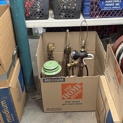 Sale Photo Thumbnail #87: Cardboard boxes filled with various vintage home goods, including disassembled table lamp parts, small light fixtures, and a collection of wall clocks.