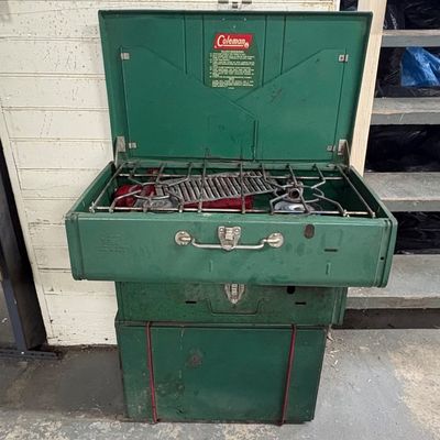 Sale Photo Thumbnail #93: Vintage green Coleman two-burner camp stove displayed open, resting on top of matching green metal storage cases.