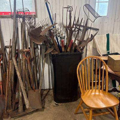 Sale Photo Thumbnail #54: Collection of vintage long-handled garden tools, shovels, pitchforks, and rakes leaning against a wall and stored in a trash bin, set next to a wooden Windsor-style chair.