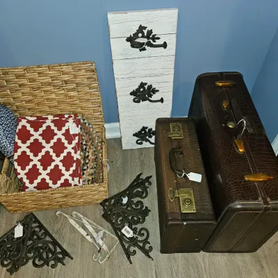 Sale Photo Thumbnail #12: Collection of home decor items including two vintage hard-shell suitcases, decorative cast iron brackets, a distressed wooden wall rack with hooks, and a wicker storage hamper filled with textiles.