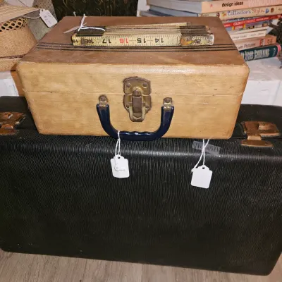 Sale Photo Thumbnail #7: Vintage small light wood-look suitcase with a dark blue handle stacked atop a larger black hard-shell suitcase, topped with a vintage folding ruler.