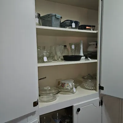 Sale Photo Thumbnail #64: White kitchen cabinet filled with various household items, including glassware, clear glass casserole dishes, a cast iron skillet, milk glass vase, and plastic storage containers.