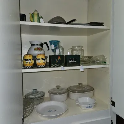 Sale Photo Thumbnail #65: A collection of vintage kitchenware stored inside a white cabinet, including CorningWare, glass casserole dishes, cow-themed salt and pepper shakers, and a teapot.