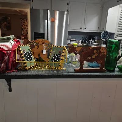 Sale Photo Thumbnail #14: Collection of country-style kitchen decor and glassware displayed on a counter, including hand-painted rooster art, a carved wooden cow figurine, green glass pitcher, and wire storage baskets.