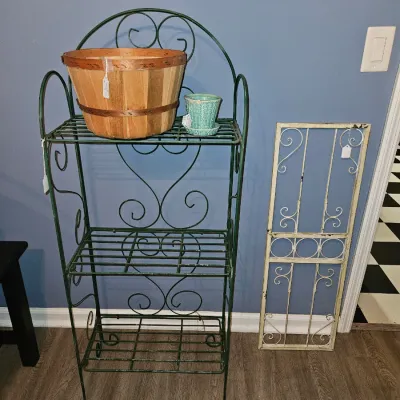 Sale Photo Thumbnail #37: Vintage green wrought iron three-tiered baker's rack holding a wood slat bushel basket and small teal planter, displayed alongside a white decorative metal trellis.