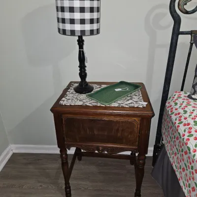Sale Photo Thumbnail #71: Vintage dark wood nightstand featuring carved details and turned legs, topped with a black table lamp with a buffalo plaid shade and a green ceramic tray.