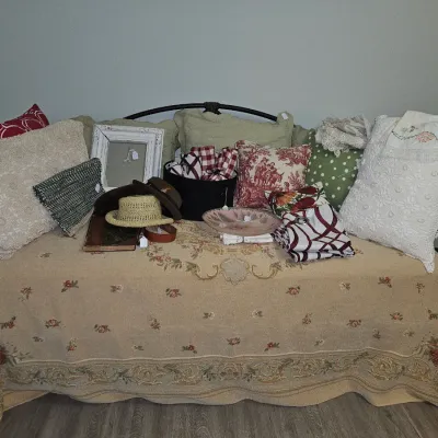 Sale Photo Thumbnail #22: Metal frame daybed dressed with an embroidered coverlet and holding an assortment of accent pillows, hats, a picture frame, and folded textiles.