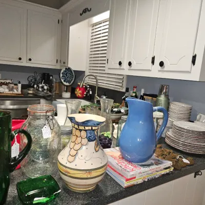 Sale Photo Thumbnail #15: Collection of kitchen glassware and ceramic items, including green glass pitchers, a hand-painted Tuscan-style vase, a periwinkle ceramic pitcher, and stacks of dishware.