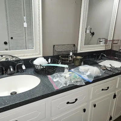 Sale Photo Thumbnail #18: Double bathroom vanity featuring white cabinetry, dark speckled countertops, framed mirrors, and various wire organization baskets and bath accessories for sale.