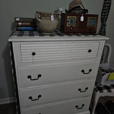 Sale Photo Thumbnail #69: White painted four-drawer dresser featuring a louvered top drawer face and dark hardware, topped with decor items including a vintage table radio and a urn-style ceramic planter.