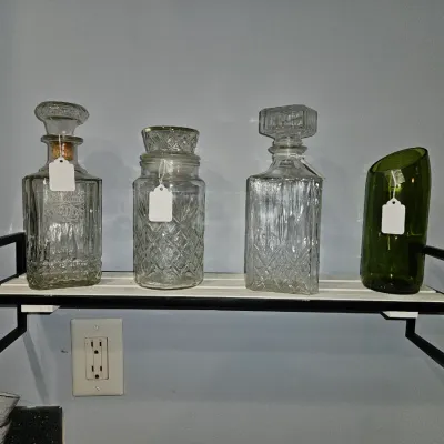 Sale Photo Thumbnail #41: Collection of glass decanters, a diamond-cut jar, and a slanted green glass vase displayed on a modern wall-mounted shelf.