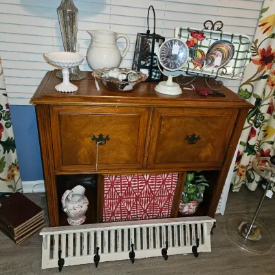 Sale Photo Thumbnail #60: Traditional wooden server cabinet featuring decorative top-tier home goods, including a rooster tray, milk glass pedestal, ceramic pitcher, and small clock, flanked by floral drapes.