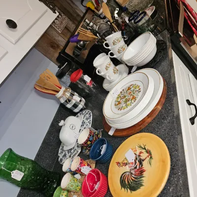 Sale Photo Thumbnail #16: Collection of vintage kitchenware and tableware on a countertop, including Corelle floral dishware, colorful ceramic mugs, a rooster plate, and wooden utensils.