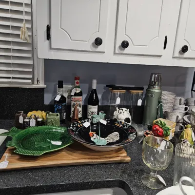 Sale Photo Thumbnail #61: Collection of kitchen items arranged on a countertop, including a green fish-shaped platter, a rooster-themed bowl, assorted glassware, vintage bottles, and a Stanley thermos.