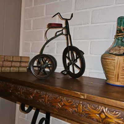 Sale Photo Thumbnail #28: Vintage miniature wrought iron tricycle decorative sculpture displayed alongside old books and a wicker-wrapped Chianti bottle on a carved wooden shelf.