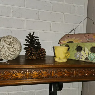 Sale Photo Thumbnail #29: Collection of decorative home accents displayed on a shelf, including a carved composite sphere, natural pine cones, a miniature yellow ceramic planter, and a painted wooden decorative birdhouse.