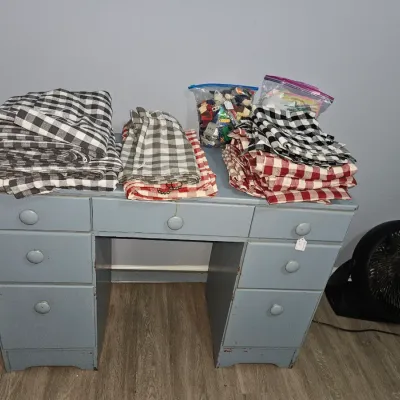 Sale Photo Thumbnail #44: Painted grey wooden desk supporting stacks of plaid and buffalo check linens and bags of craft supplies.