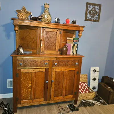 Sale Photo Thumbnail #11: Vintage oak hutch with carved details and upper storage cabinet, displayed alongside decorative home accents, luggage, and wall art.