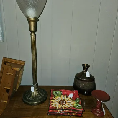 Sale Photo Thumbnail #26: Tall brass torchier table lamp with a frosted glass shade, displayed alongside decorative sunflower plates, a metallic vase, and a small red pillar candlestick holder.
