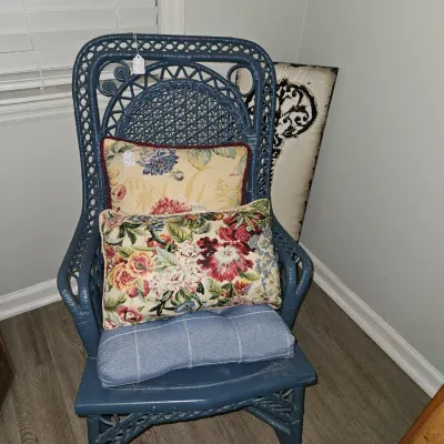 Sale Photo Thumbnail #47: Blue painted wicker rocking chair with ornate woven back detailing, fitted with a blue seat cushion and two floral throw pillows.