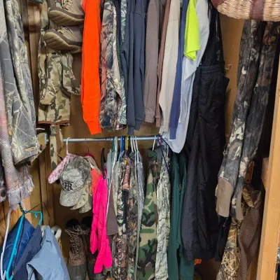 Sale Photo Thumbnail #75: Selection of outdoor and hunting apparel, including camouflage jackets, high-visibility tops, overalls, and work boots hanging inside a wooden wardrobe.