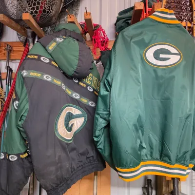 Sale Photo Thumbnail #70: Two vintage Green Bay Packers winter jackets hanging in a storage area, including a hooded heavyweight parka and a classic satin bomber jacket.