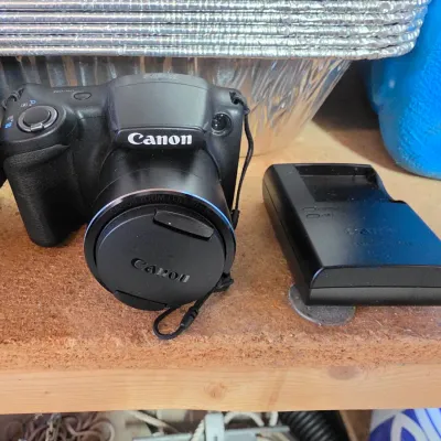 Sale Photo Thumbnail #43: Black Canon PowerShot digital bridge camera with a lens cap and a matching battery charger.