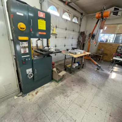 Sale Photo Thumbnail #91: Large industrial vertical band saw machine alongside a mobile rolling work cart and an orange shop crane hoist in a workshop.