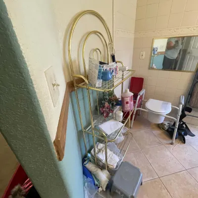 Sale Photo Thumbnail #8: Brass-toned tubular metal baker's rack featuring arched top details and wire shelves, displayed alongside bathroom supplies and a bedside commode chair.