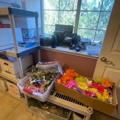 Sale Photo Thumbnail #82: Craft supplies and artificial flowers arranged on plastic shelving units near a window, alongside a boombox and electronics on the sill.