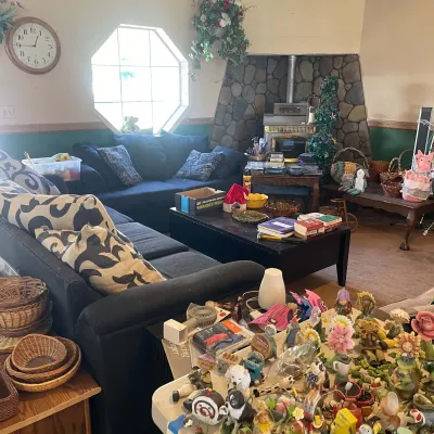 Sale Photo Thumbnail #57: Living room filled with household items, including a dark upholstered sectional couch, coffee table, stone fireplace, and a table loaded with colorful knick-knacks and figurines.