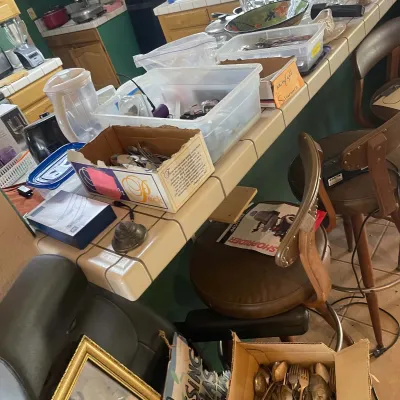 Sale Photo Thumbnail #62: Kitchen counter and bar stools laden with assorted household goods, plastic storage bins, silverware, and kitchenware.