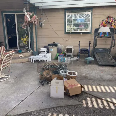 Sale Photo Thumbnail #79: Outdoor collection of home goods and exercise equipment including a treadmill, blue rollator walker, pet crates, patio chairs, and cardboard boxes with various household items.