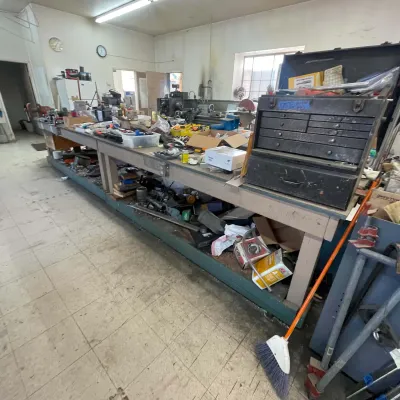 Sale Photo Thumbnail #101: Long industrial workshop workbench cluttered with various tools, hardware, bench vises, and a metal machinist tool chest.