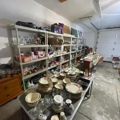 Sale Photo Thumbnail #29: Garage estate sale setup featuring folding tables stacked with floral china dishes, glassware, and decorative items, alongside plastic shelving units filled with household goods.