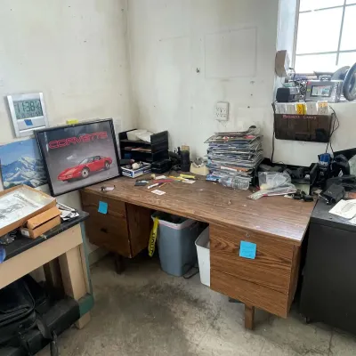 Sale Photo Thumbnail #39: Vintage wood laminate office desk with drawers, featuring a framed red Corvette poster, office supplies, and stacked magazines.